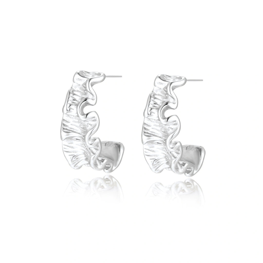 Textured white gold-plated hoop earrings with flowing wave pattern against cream background, showing dimensional rippled surface.