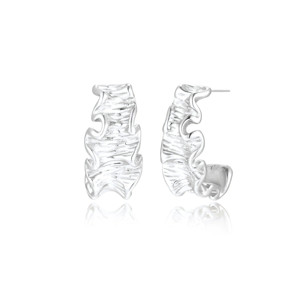 Textured white gold-plated hoop earrings with flowing wave pattern against cream background, showing dimensional rippled surface.