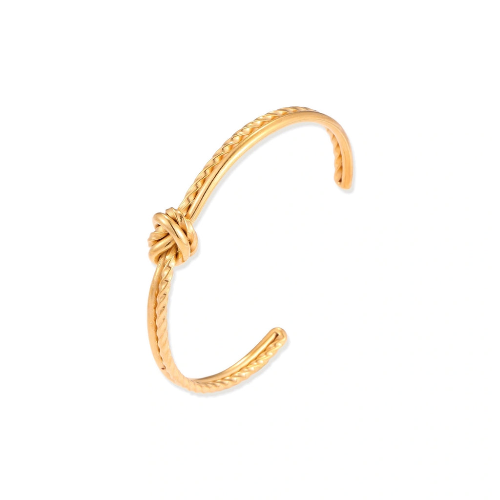 Gold cuff bracelet with twisted rope texture and decorative knot centerpiece on cream background.